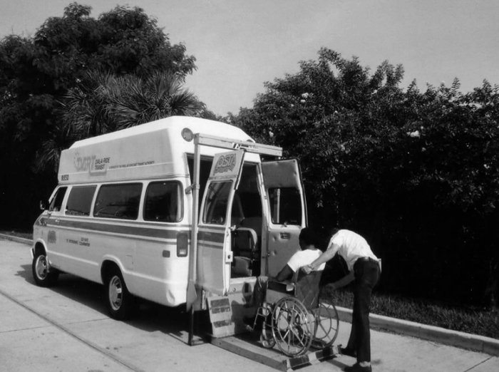 A black and white photo of a DART paratransit operating assisting a customer with a mobility aid (wheelchair) into a DART Dial-A-Ride Van. This image comes from when PSTA took over DART, but it's similar to when SPMTS operated the service.