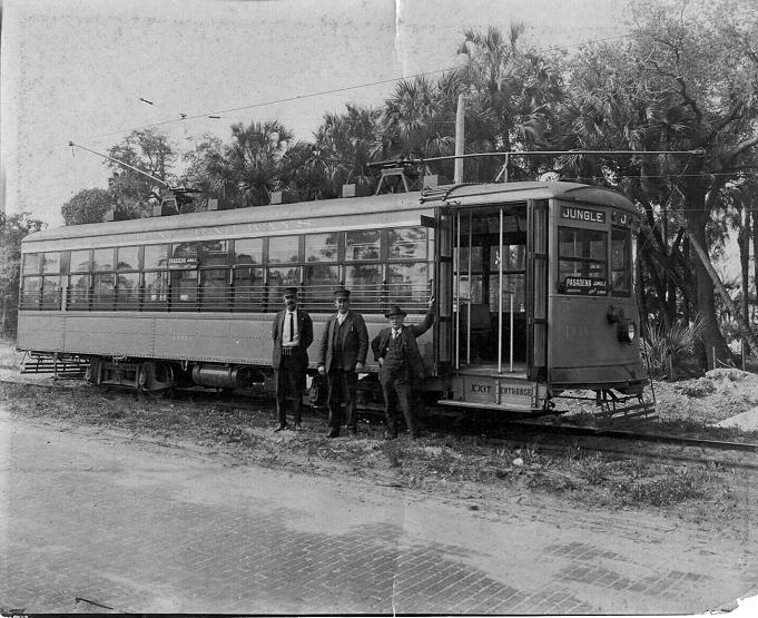 An old black and white photograph of the Jungle Line Trolley. This trolley has enclosed windows and three employees or motormen are standing outside the front door to the trolley.