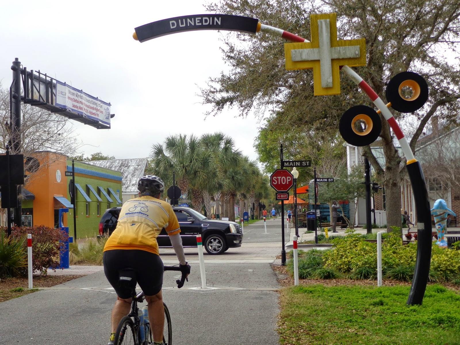 A person riding a bicycle on the Pinellas Trail underneath a curved, railroad-inspired sign that says "Dunedin."