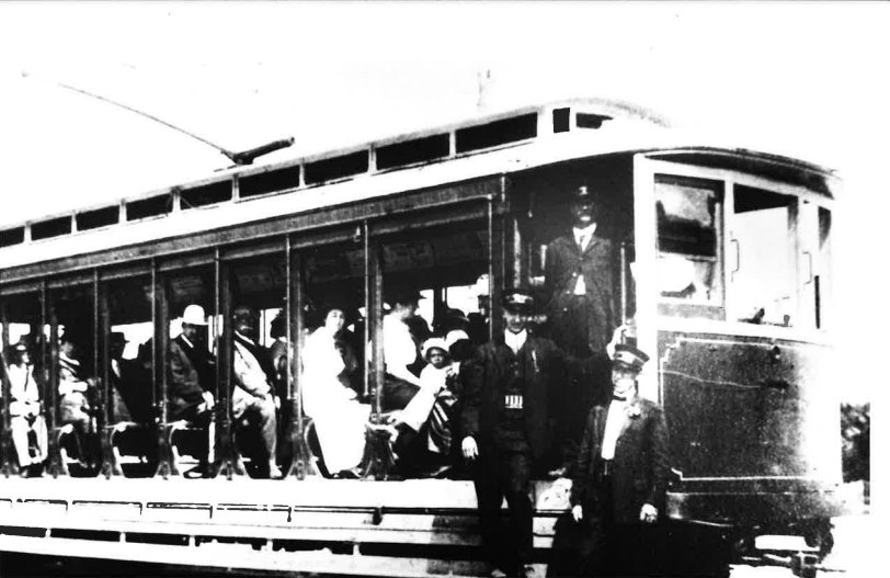 An old black and white photograph of a a trolley in 1919. Passengers sit in the open-air seats and three employees or motormen stand by the front.