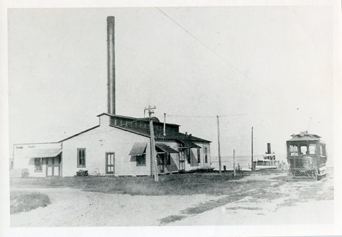 A black and white picture of streetcar #1 at the start of Central Avenue on Tampa Bay. Next to the streetcar is a white building with a tall smoke stack: this is the powerhouse for the electric streetcar system.