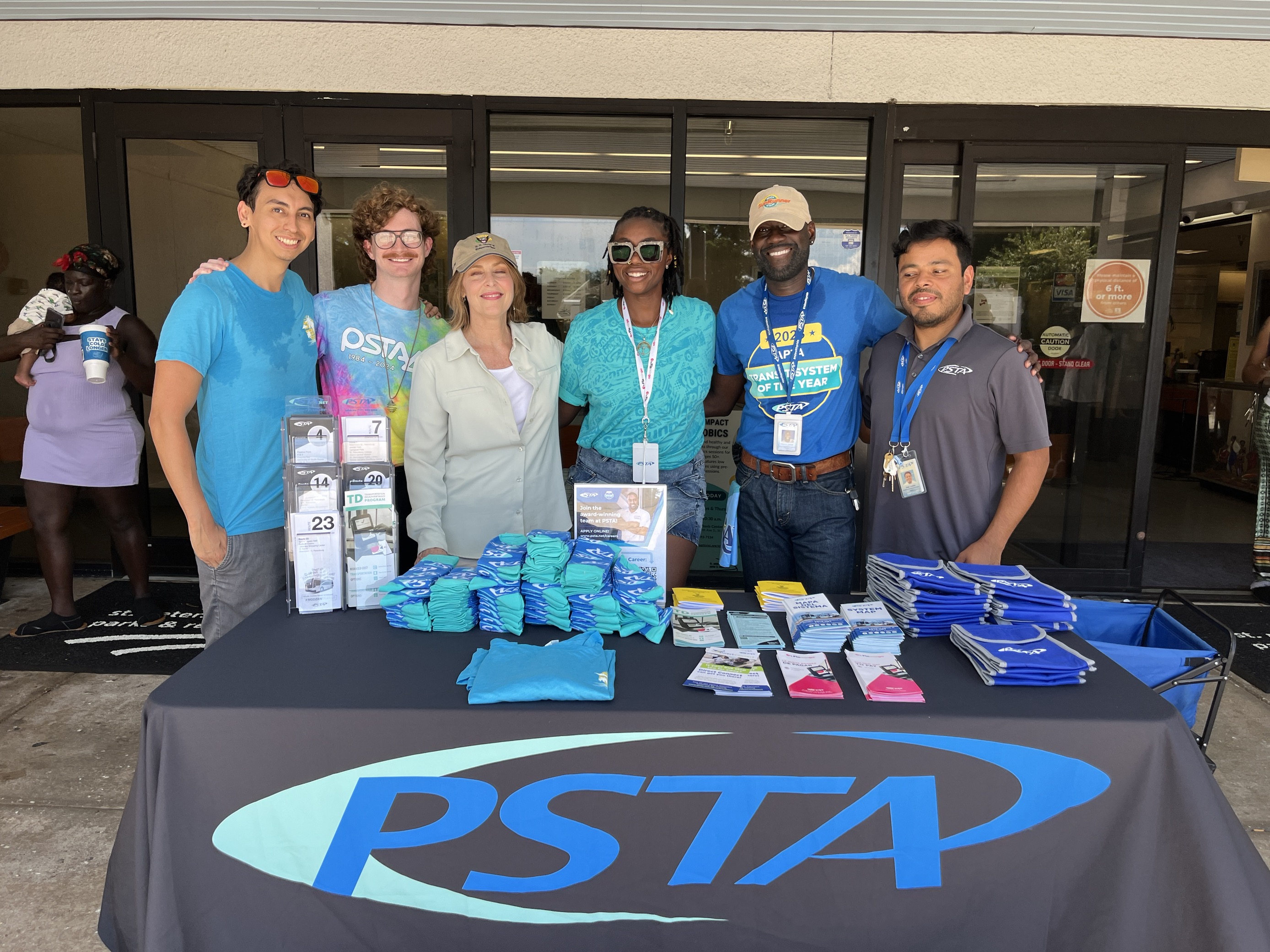 Representative Kathy Castor joins PSTA's Marketing and Outreach team at a hurricane relief drive in partnership with Reach St. Pete. Six people stand behind a tabled labels PSTA where supplies and informational brochures about taking transit are being handed out to people who lost their vehicles and homes to Hurricane Helene.