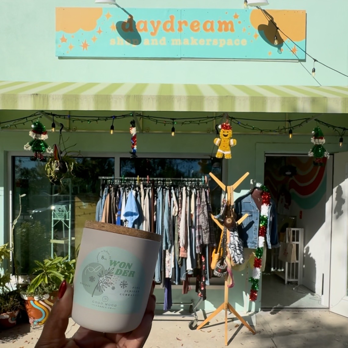 A photo of the front of Daydream Shop and Makerspace. Someone is holding a candle in front of the shop and in the background you can see a rack of clothes and the minty blue and yellow sign with the business name.