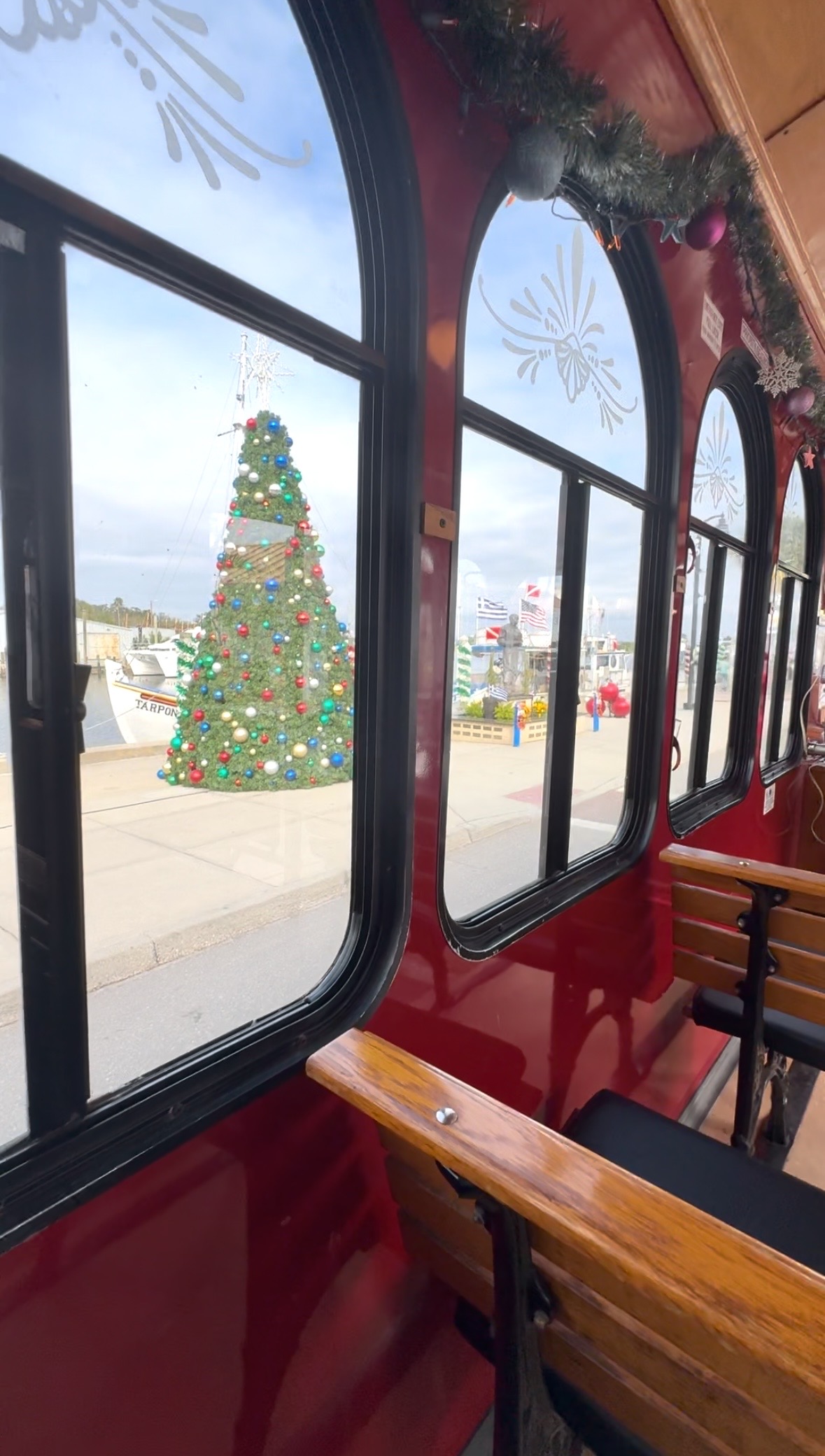 A photo looking out the window of a red and yellow trolley, where you can see a holiday tree on the sidewalk of the Sponge Docks in Tarpon Springs