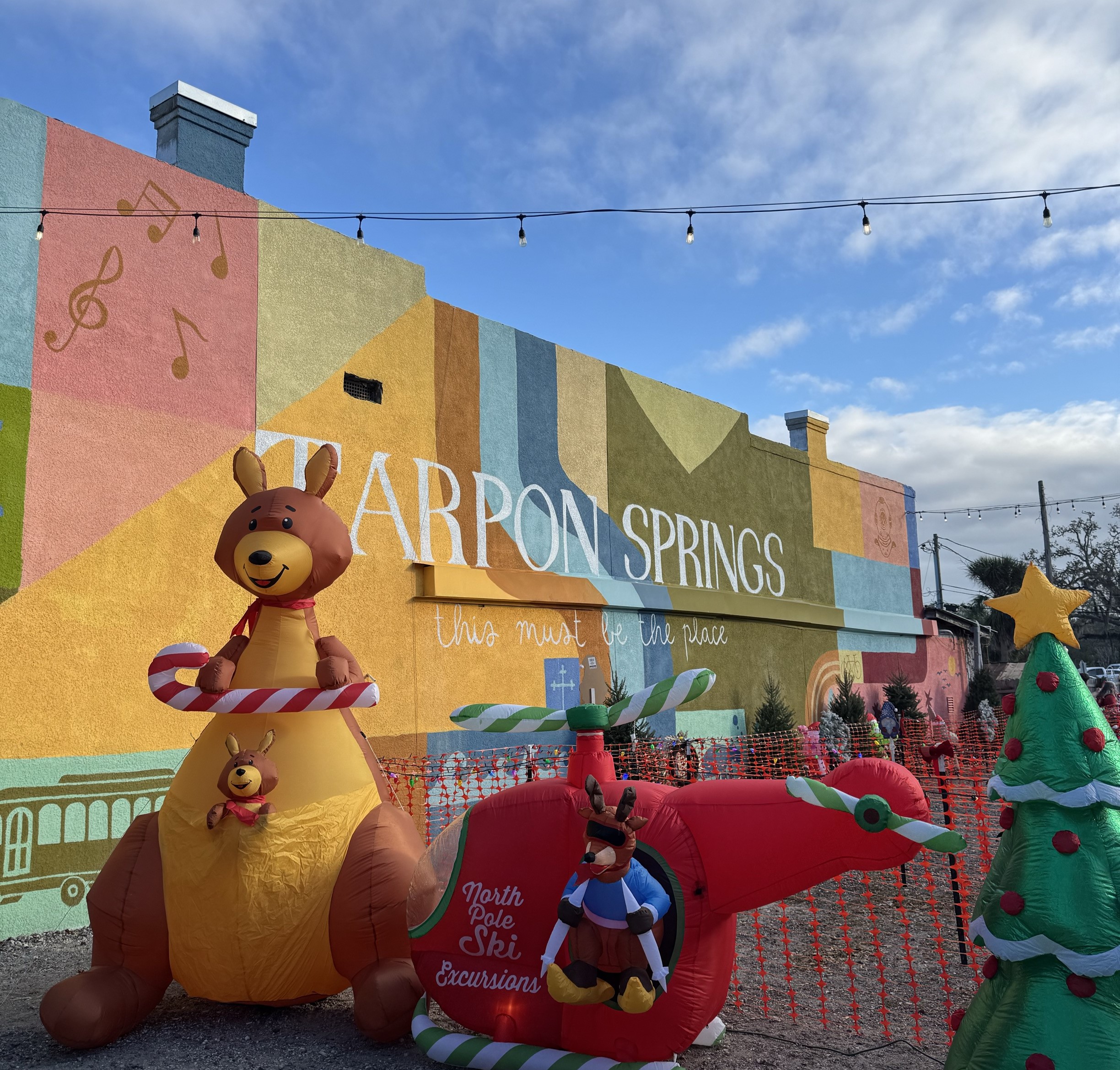 A photo of a holiday display in Tarpon Springs, featuring an inflatable kangaroo, holiday tree, and a skiing reindeer coming out of a helicopter. Behind the display is a colorful mural that says "Tarpon Springs, this must be the place"