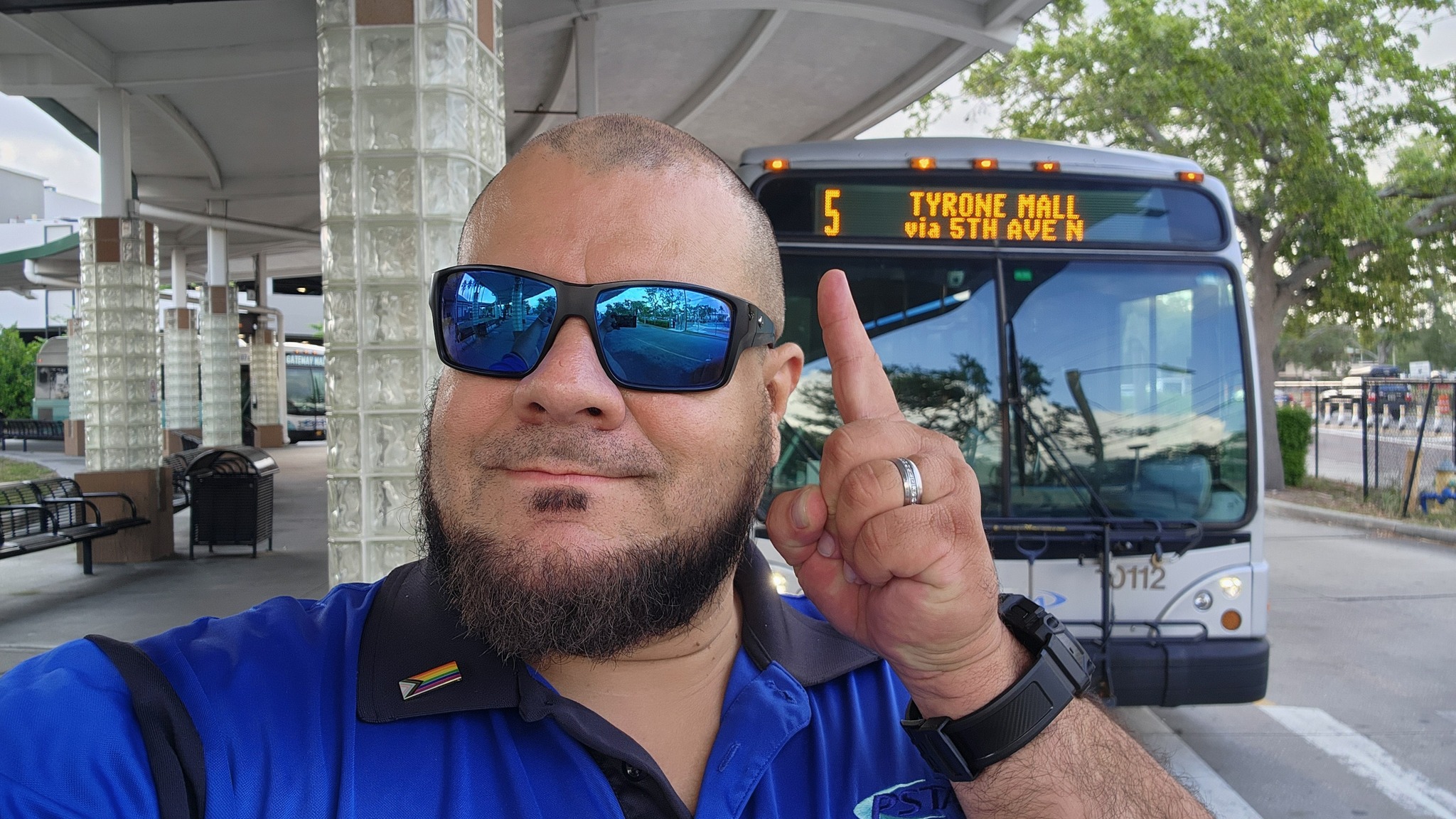Brian smiles while standing in front of a bus, pointing to the Route 5 label on the headsign. He's wearing a black and blue PSTA shirt and sunglasses.