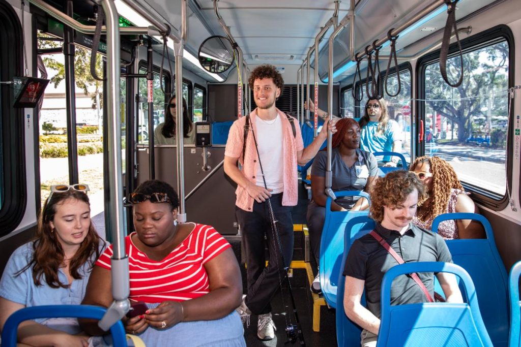 man standing on bus with fishing pole while other riders chat.