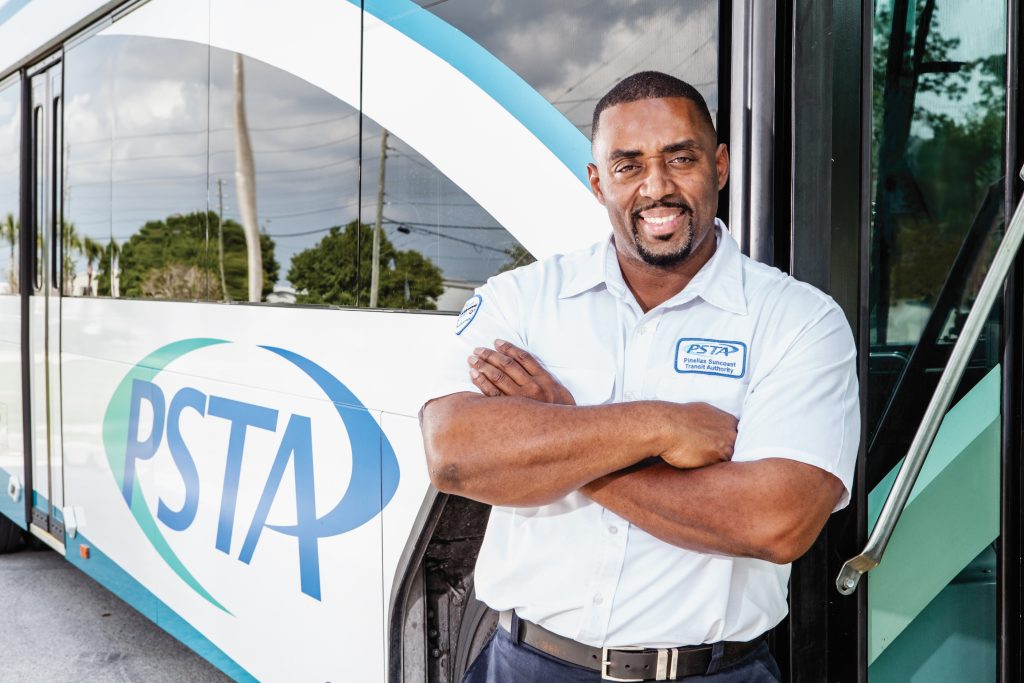 A photo of a PSTA bus operator standing at the entrance of a PSTA bus. He is smiling and has his arms crossed.