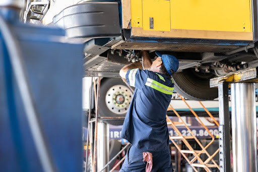 A technician reaching up into the underbelly of a lifted bus to perform maintenance.
