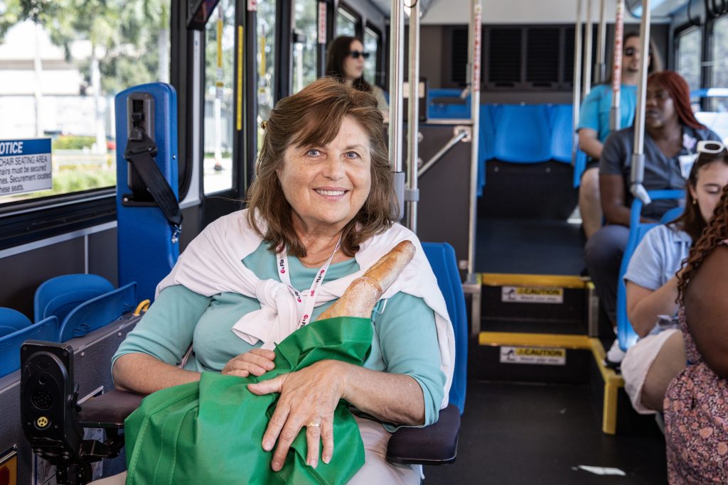A photo of Gloria seated and smiling in the wheelchair section of a bus, holding a bag of groceries in her lap.