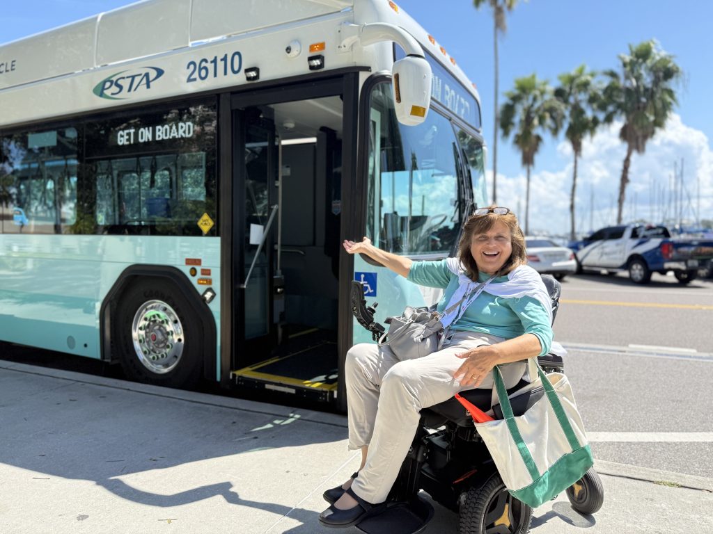A photo of Gloria in front of a bus, gesturing towards the open door with a big smile.