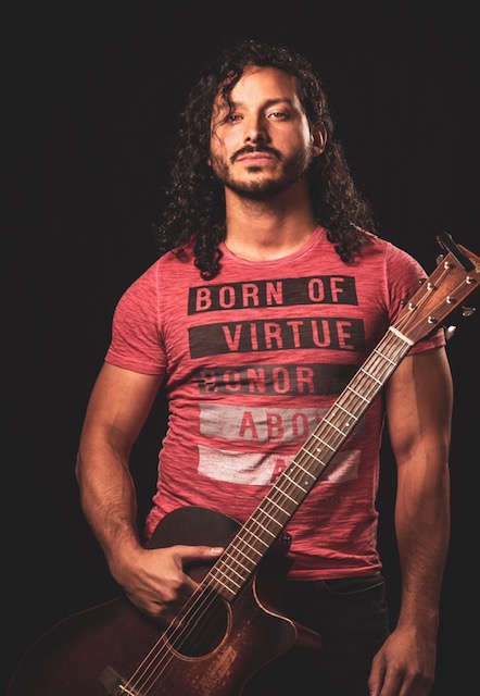 Photo of a man with long hair holding a guitar and looking seriously at the camera.