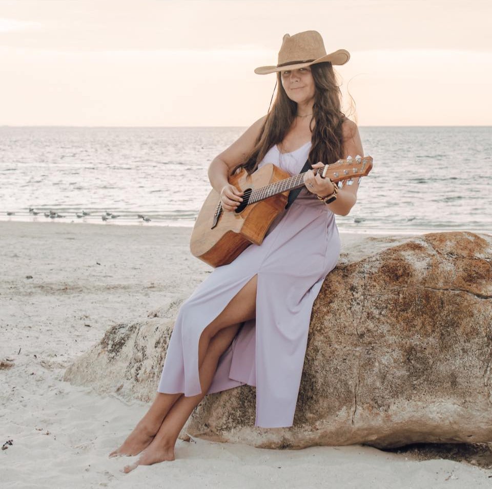 A woman wearing a hat and playing a guitar on the beach at sunset: this is Mallory Moyer