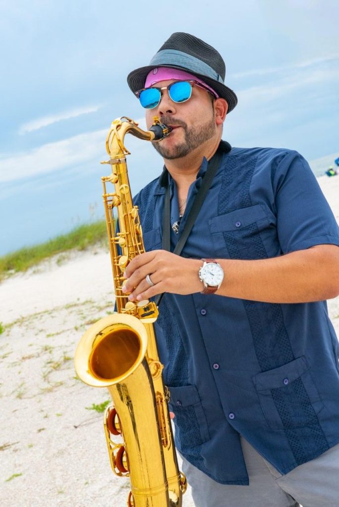 Man wearing a hat and sunglasses playing sax on the beach: this is Omar Lopez