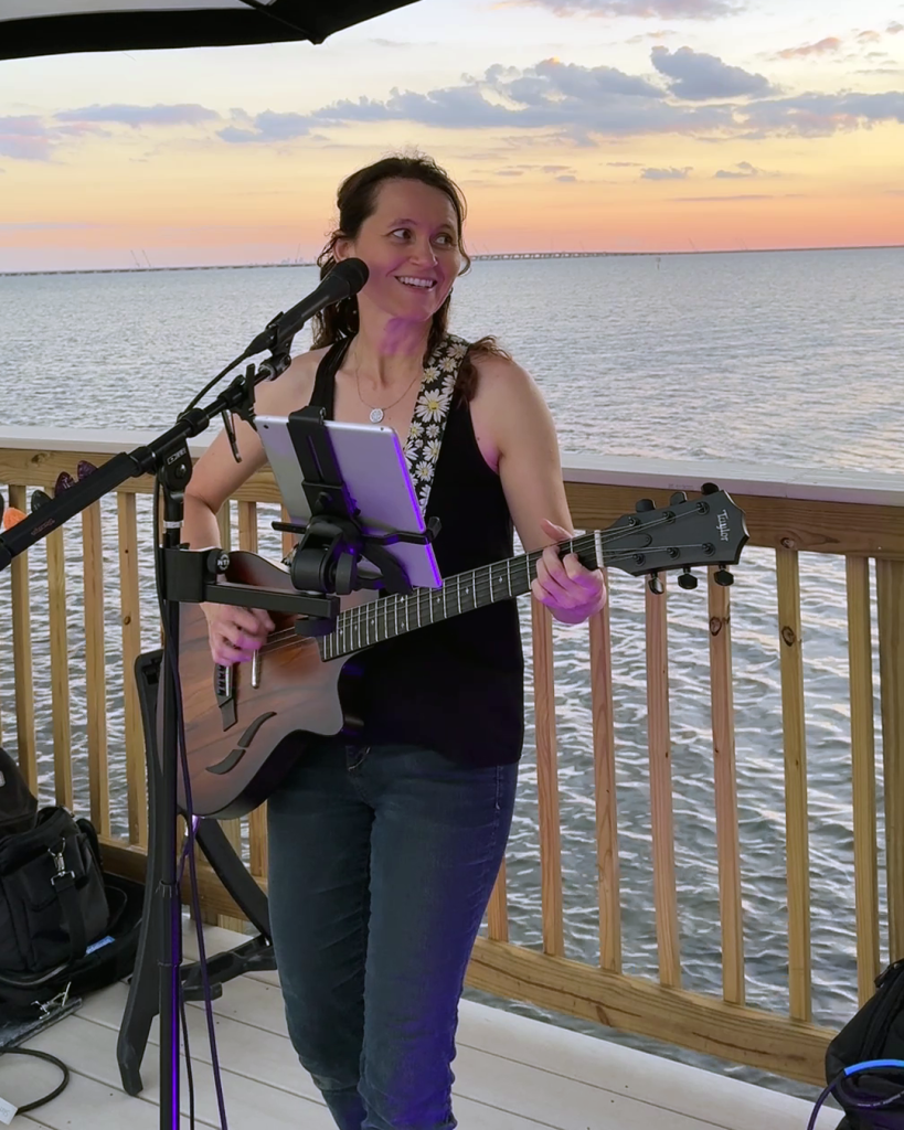 A woman playing guitar near the water with the sunset behind her. This is Susan B.